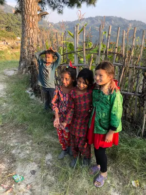 Four children, two girls and two boys, stand in a grassy area with a tree and a bamboo fence in the background.