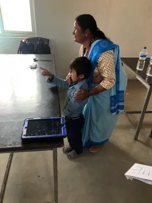 A woman and a boy are standing in front of a table with a tablet on it.