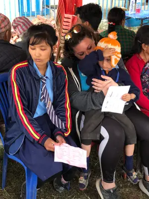 A girl with a school uniform reads a piece of paper while sitting on a chair while an adult holds a young child on her lap and also reads a piece of paper