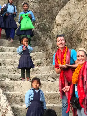 A group of people, including young girls, are standing on stone stairs and smiling for a photo, one of them carrying a green bag