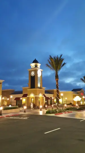 A large clock tower with a clock face and a palm tree in front of a shopping mall at night