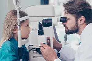 A doctor is examining a young girl's eyes with a microscope in an examination room.