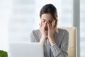 woman closing her eyes while sitting at a desk with her hands on her face
