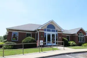 A brick building with glass windows and a glass door in front of a lawn with plants and bushes, a metal railing on the side, and a parking lot in front under a blue sky.