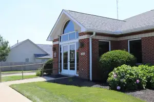 A brick building with glass doors and windows, a gray roof, and a garden with bushes and grass in front of it, with a house and fence in the background.