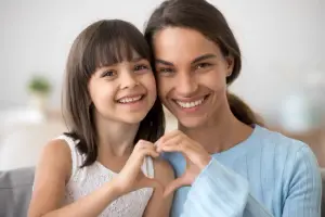 A woman and a young girl sitting together and making a heart shape with their hands while smiling