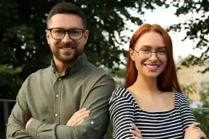 A man and a woman standing together in a park, both wearing glasses and smiling