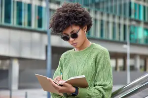 A woman in a green sweater and sunglasses writes in a notebook on an escalator.