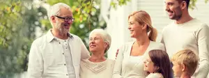 A family is smiling in front of a house, the man on the left has glasses and the woman next to him has white hair, a woman with a ponytail next to her and a child with blonde hair in front of her.