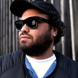 A man with a beard wearing a black hat and sunglasses is standing outside in front of a wall.