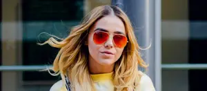 A woman with blonde hair wearing sunglasses and a yellow shirt is standing in front of a building.