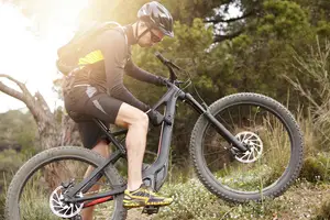 A person is riding a mountain bike on a grassy trail in the woods with sunlight illuminating the scene.