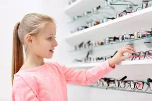 A young girl in a pink shirt is choosing glasses from a store shelf.