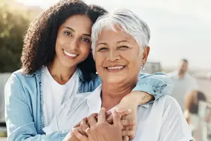 A woman is hugging an elderly woman and smiling while posing for a photo. They are both wearing white shirts, and the elderly woman has gray hair. Behind them, a person is standing, and a building is in the distance. There are plants and trees in the area, and the sky is covered with clouds.