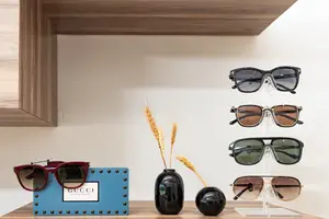 A wooden shelf displays several pairs of sunglasses, including Gucci and Tom Ford brands, along with decorative items such as a blue sign, a wheat stalk in a vase, and a red pair of sunglasses on top of a blue box.