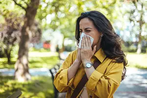 A woman in a yellow jacket is blowing her nose in a park.