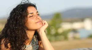 A woman with curly hair and a smiling face sitting on a bench, with her eyes closed and her hand resting on her chin, while a tree and a building are in the background