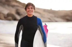 A man in a black wetsuit is smiling and holding a surfboard on the beach near the sea.