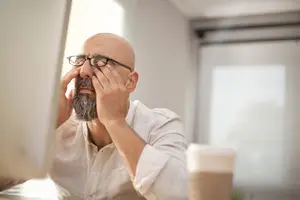 Man in glasses sitting at desk with hands on face rubbing eyes
