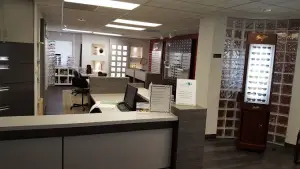 Interior view of an optometrist office featuring a front desk, a reception area with a monitor, chairs, and a glass wall with sunglasses on display, all under ceiling lights.