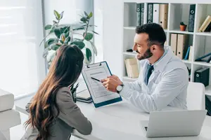 A man in a white coat is talking to a woman in a suit who is sitting at a desk in an office.