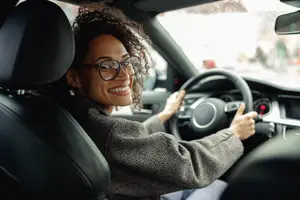 A smiling woman with glasses driving a car