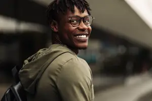A smiling man wearing a green jacket, glasses, and a backpack, standing on a street near a building with a blurred background