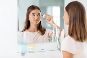 A woman uses a bottle to apply something to her eye in front of a mirror