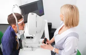 A young boy is undergoing an eye examination by a woman in a white uniform using a monitor and eye scanner.