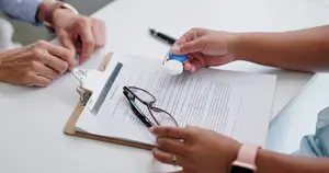 Two people sitting at a table with a clipboard, one holding a pair of glasses and the other holding a blue cap.