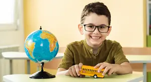 A young boy with glasses holds a toy bus and smiles at a globe in a classroom.