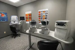 An empty room with a table, chairs, and medical equipment. The room has gray walls, a ceiling light, and a poster on the left wall. The table has a Nikon machine, a monitor, and a black chair. The chairs are black, and the floor has a gray carpet.