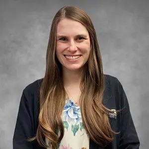 A smiling woman with long brown hair wearing a black jacket and floral dress standing against a gray background