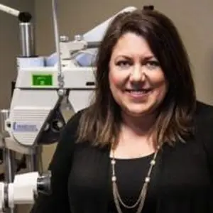A woman wearing a black top and necklace is smiling for a picture while standing in a room with a medical machine behind her.