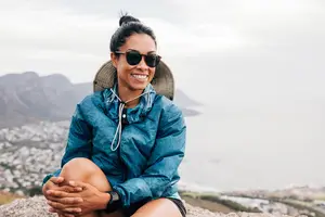 A woman wearing a blue jacket, sunglasses, and a hat is sitting on a rock and smiling while looking at the camera. Behind her, there is a view of a mountain and a city.