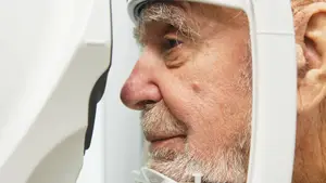 An older man wearing glasses is looking through a white eye exam device in a room