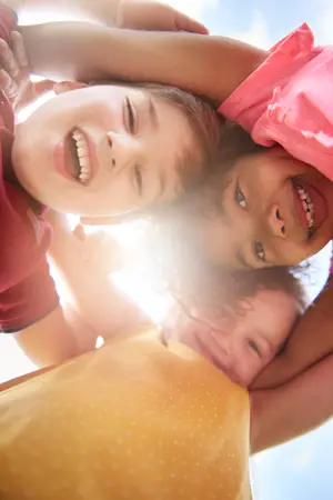 Three young children, one boy and two girls, are smiling and appear to be posing for a photograph with their heads together in a circle, and the sunlight is shining on them.