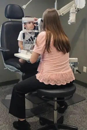 A young girl sits in a black chair examining a child's eyes through a white machine in a medical office