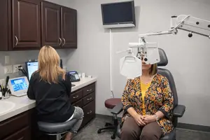 A woman with long hair is sitting on a chair in front of a monitor in a room with cabinets and a sink. She is wearing a yellow top and brown pants. A woman with blonde hair is sitting on a chair in front of a monitor and a television mounted on the wall. She is wearing a black top and white pants. There is a telephone on the desk and a sink with a faucet on the left.