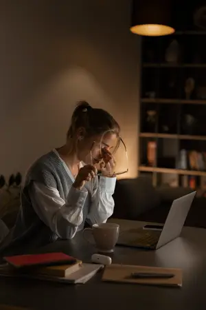 A woman sitting at a table with a laptop, a cup, books, and a pen, possibly in a study room with a lit lamp and a shelf with books and vases