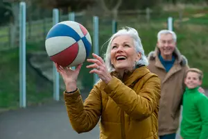 An older woman holds a basketball in an outdoor sports court, smiling while an older man and a child stand behind her, all wearing jackets and smiling