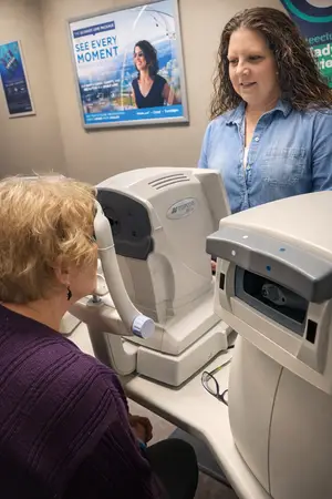 An adult woman with blonde hair is using a Topcon machine to check her eye sight while another woman is standing behind her.
