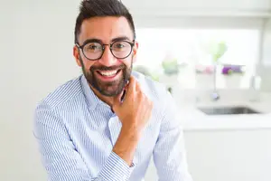 A smiling man in a blue striped shirt is adjusting his glasses while looking sideways in a kitchen setting.