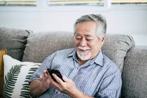 A man with white hair and beard sitting on a couch and holding a cell phone