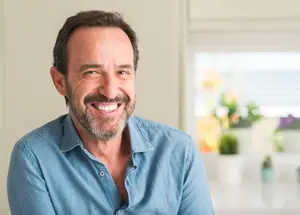 A smiling man with a beard and mustache stands in a room with a white wall and a blurry view of plants in the background.