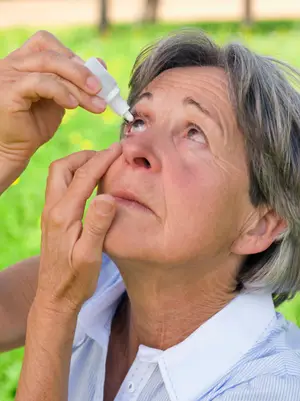 An older woman is applying eye drops to her eye on a grassy field with a blurry background.