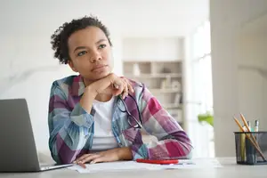 A young woman sitting at a desk in a home office wearing glasses and a plaid shirt, holding her chin with her left hand, looking thoughtfully at a laptop on the desk and papers, a basket of pencils, and a red pen.