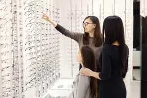 Three women are looking at a variety of eyeglasses displayed on a wall in a store