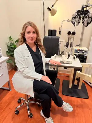 A woman wearing a white coat and white sneakers is sitting on a chair in an eye clinic, smiling and posing for a photo.