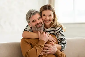A woman hugging a man who is sitting on a couch and smiling
